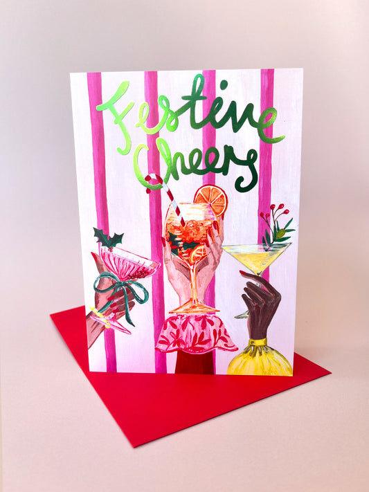 A festive greeting card with the words 'Festive Cheers' in green script over pink vertical stripes, featuring illustrated cocktails, shown standing on a red envelope against a light beige background.