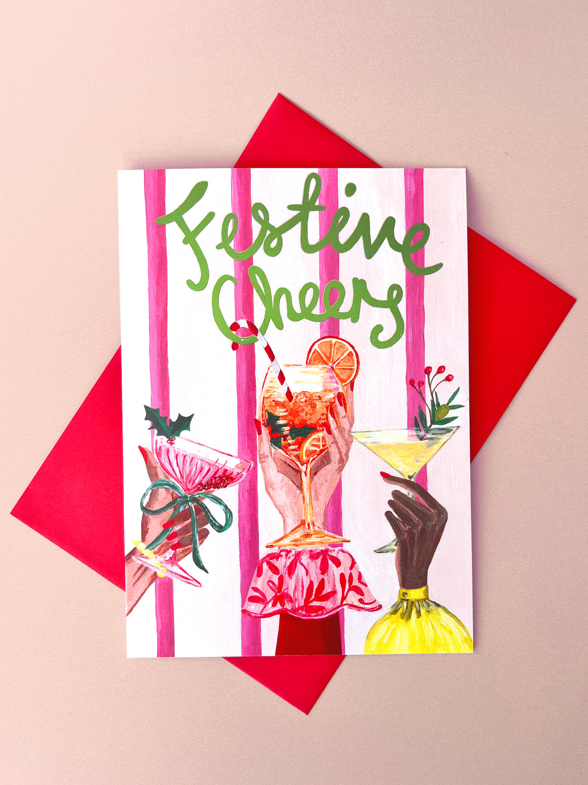 A festive greeting card with pink vertical stripes and the words 'Festive Cheers' in green script, shown with a red envelope on a beige background. The card features illustrated cocktail glasses and hands holding drinks.