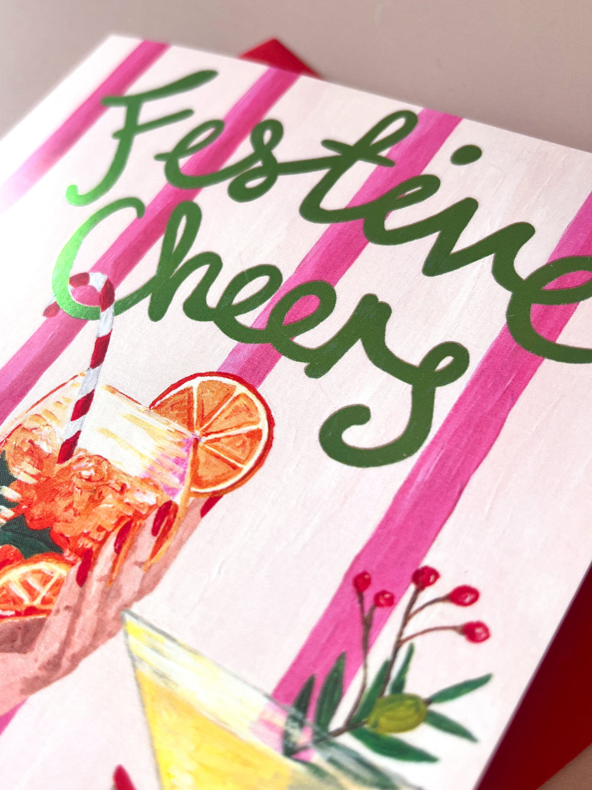 A close-up of a printed card or box cover with the words 'Festive Cheers' in green script over pink stripes, decorated with illustrations of a cocktail, orange slices, and small red berries.