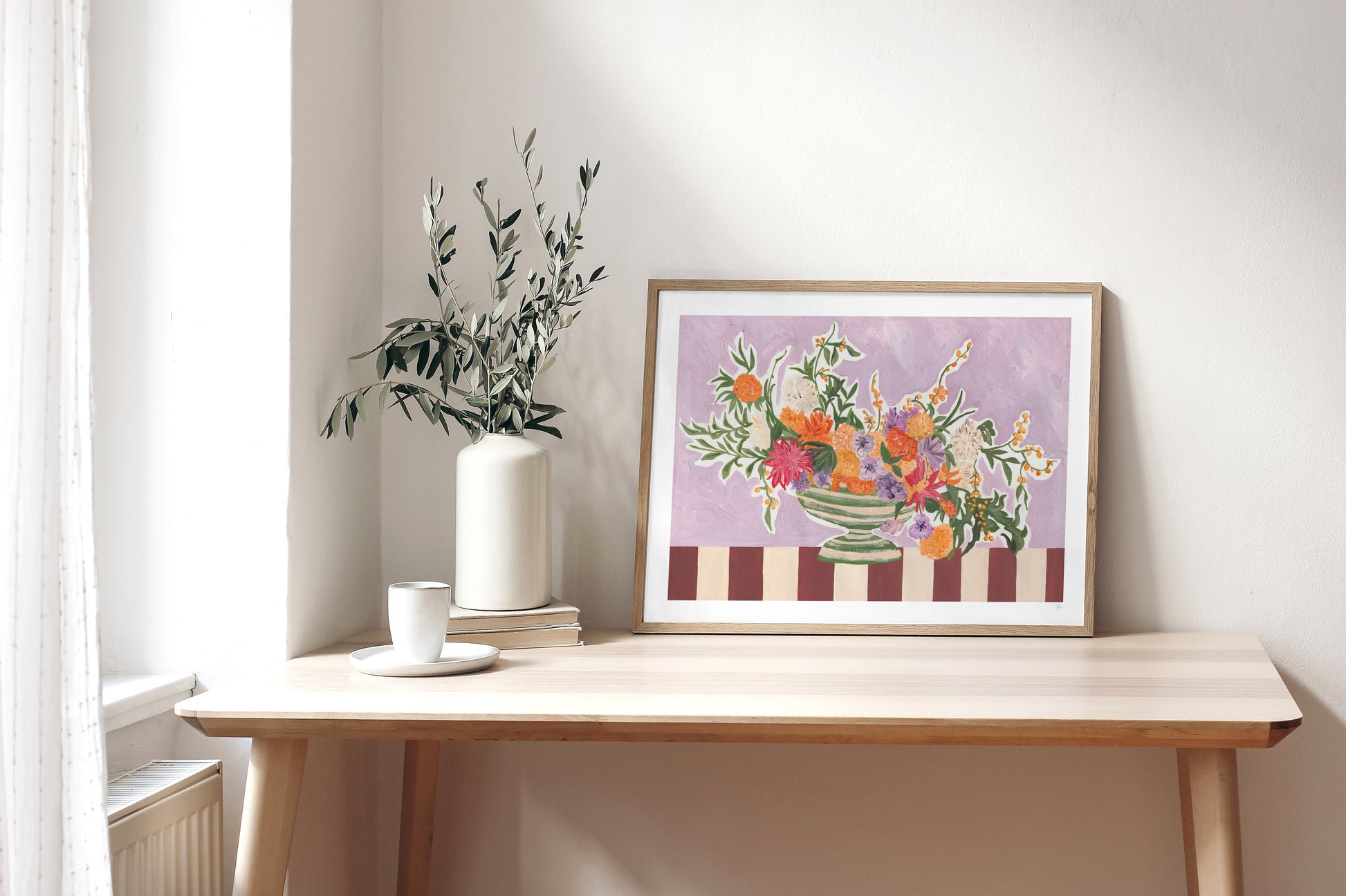 Framed floral artwork on a wooden table with a vase of flowers and a cup.