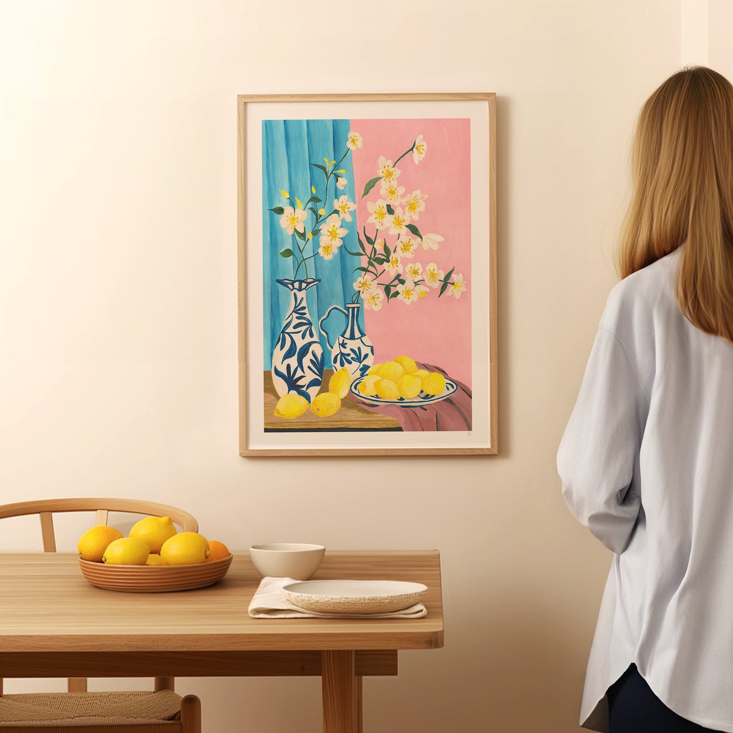 Woman looking at a framed painting of lemons and flowers on a wall above a table with lemons.