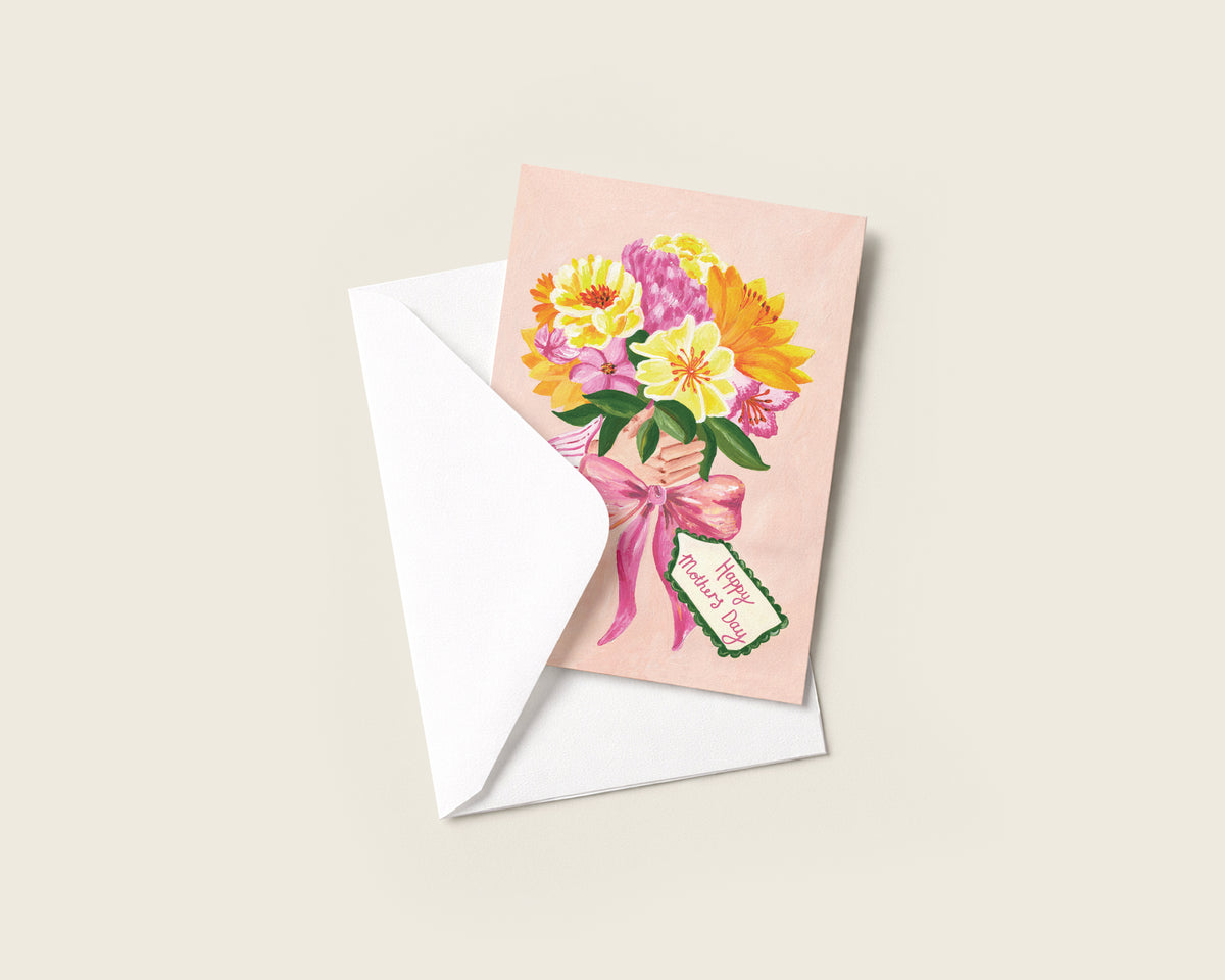 A square greeting card with a pastel pink background and a colorful bouquet illustration in yellow, pink, and orange, shown with a white envelope on a light beige background.
