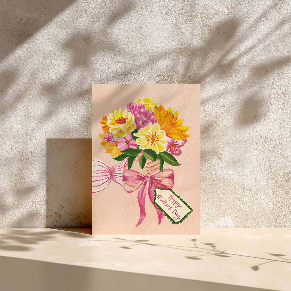 A Mother's Day greeting card featuring a colorful watercolor bouquet of yellow, pink, and white flowers tied with a pink ribbon and a small tag reading 'Happy Mother's Day,' displayed on a beige surface against a textured wall with leaf shadows.