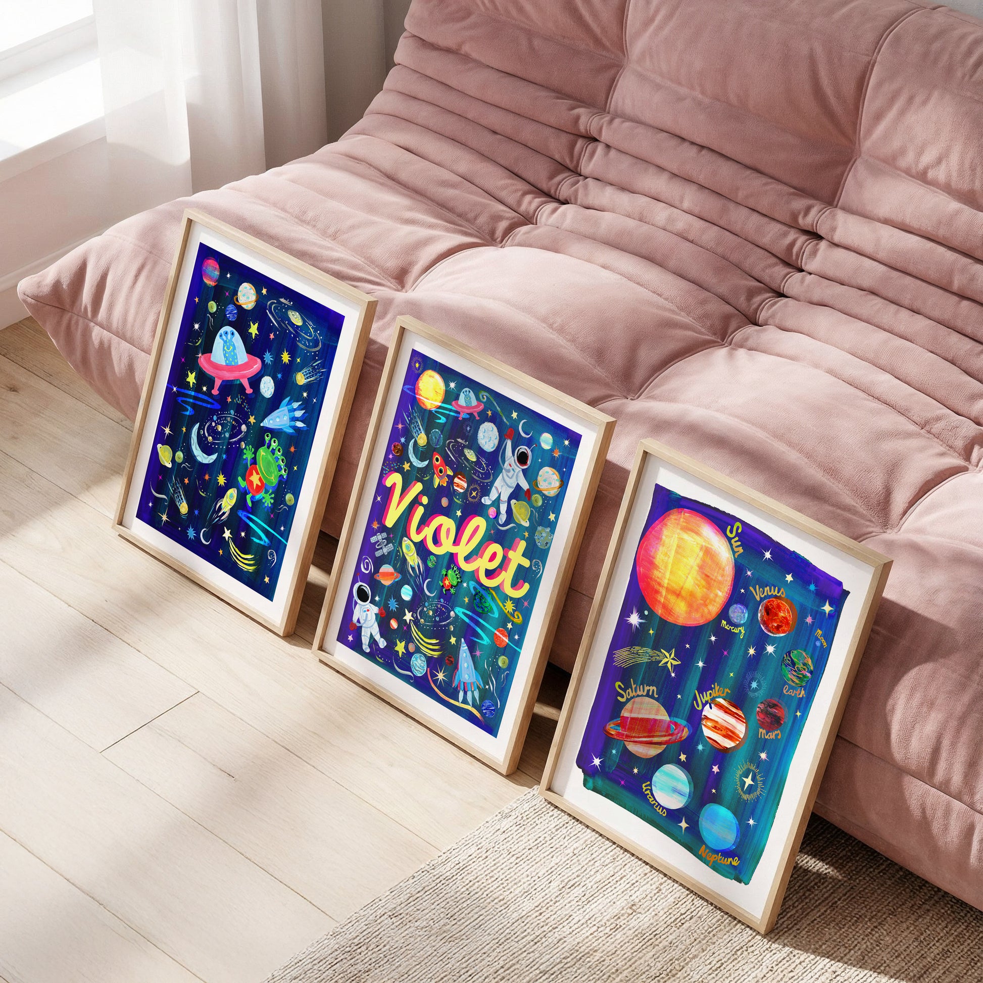 Three colorful framed artworks on a wooden floor with a pink sofa in the background.