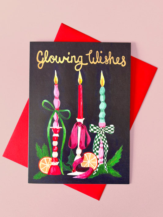 A square greeting card with a dark background and gold script reading 'Glowing Wishes,' decorated with three colorful candles, green leaves, and orange slices, placed on top of a red envelope against a pale pink background.