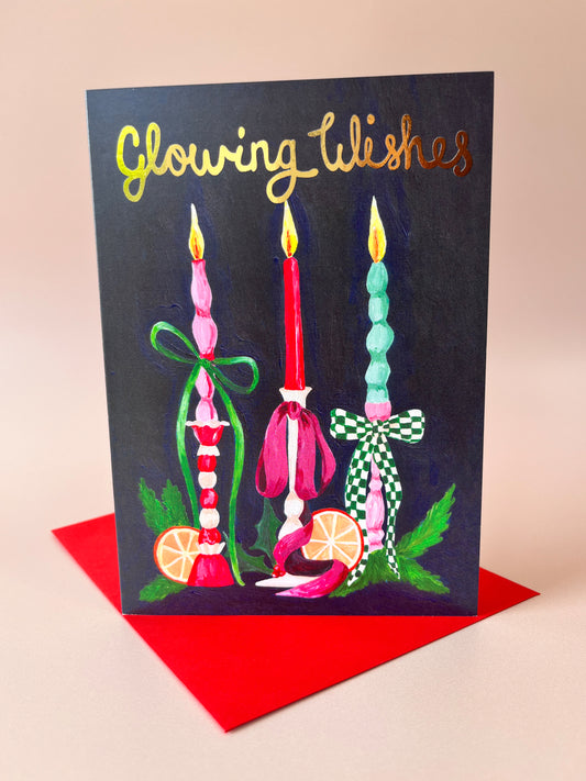 A greeting card with a dark navy background and gold foil text reading 'Glowing Wishes,' featuring three decorative candles in pink, red, and mint green with green leaves and orange slices. The card is shown standing upright on a red envelope against a warm beige background.