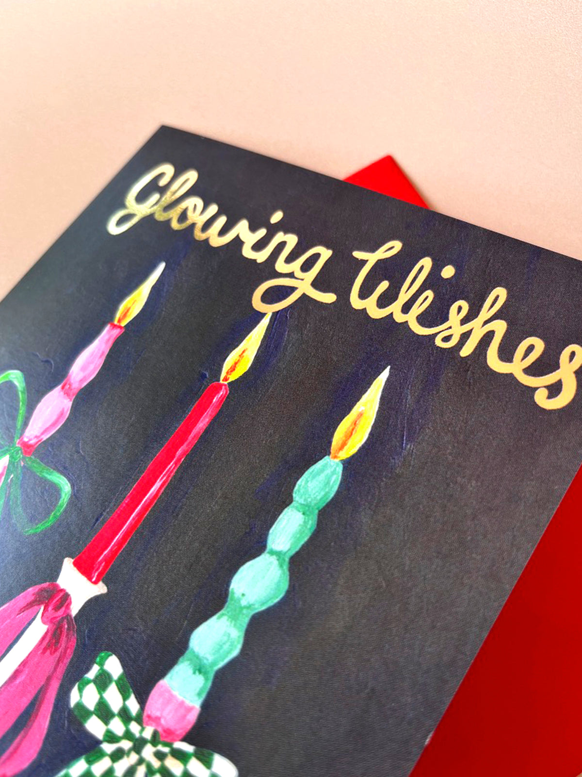 A close-up of a greeting card with the words 'Glowing Wishes' in gold script on a dark background, decorated with colorful illustrated candles, with a red envelope partially visible behind it.