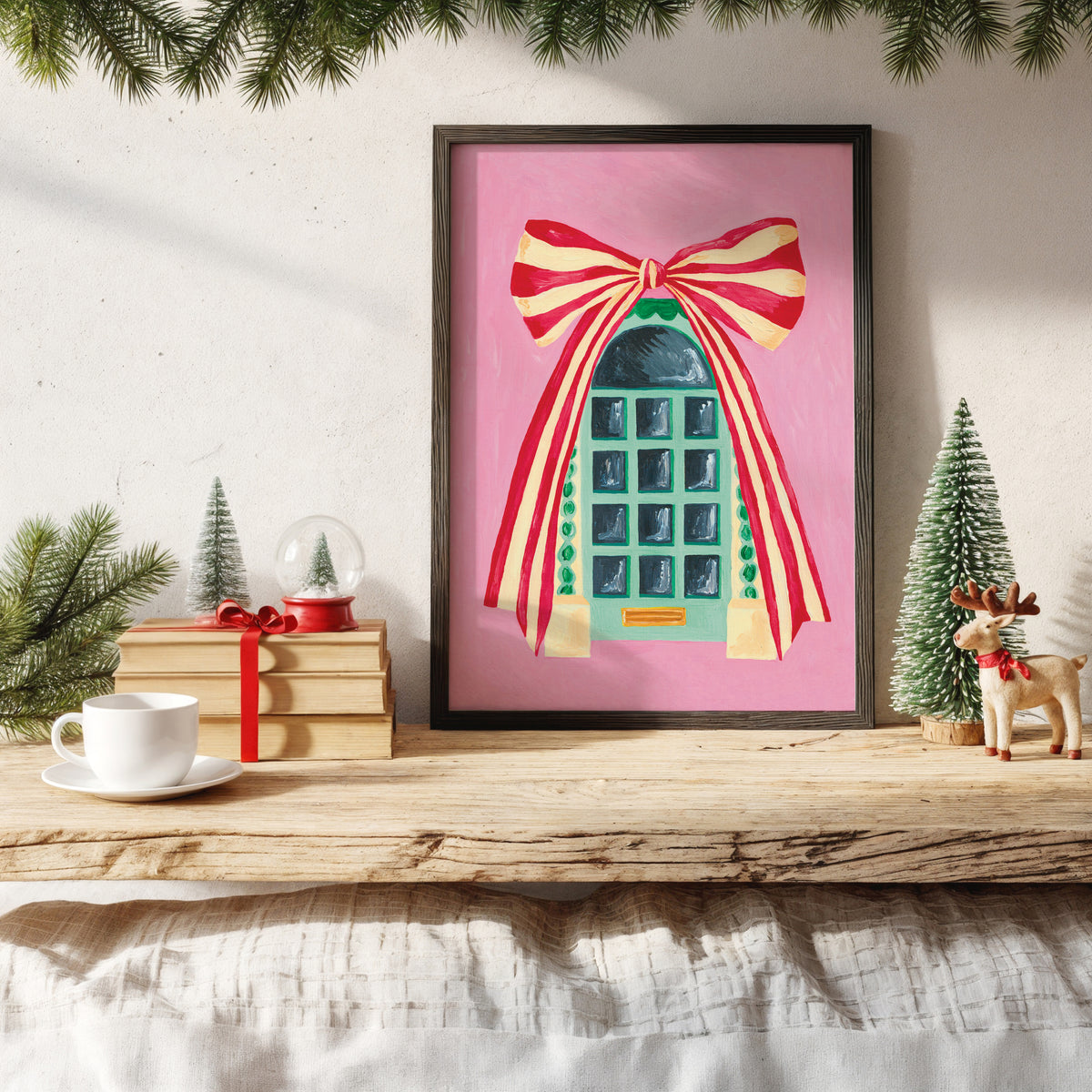 A framed art print featuring a whimsical teal door with a large red and yellow striped bow, displayed on a rustic wooden shelf in a styled holiday setting with a white cup, gift box, and small trees.