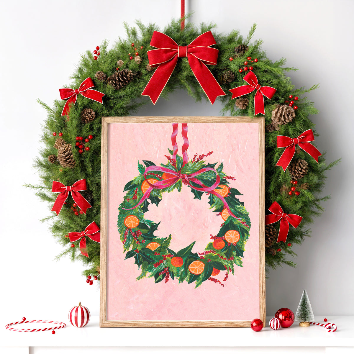 A framed Christmas art print featuring a festive wreath with red ribbon bows, green holly leaves, pinecones, and orange slices on a pink background, styled on a white surface with holiday ornaments and a small tree.