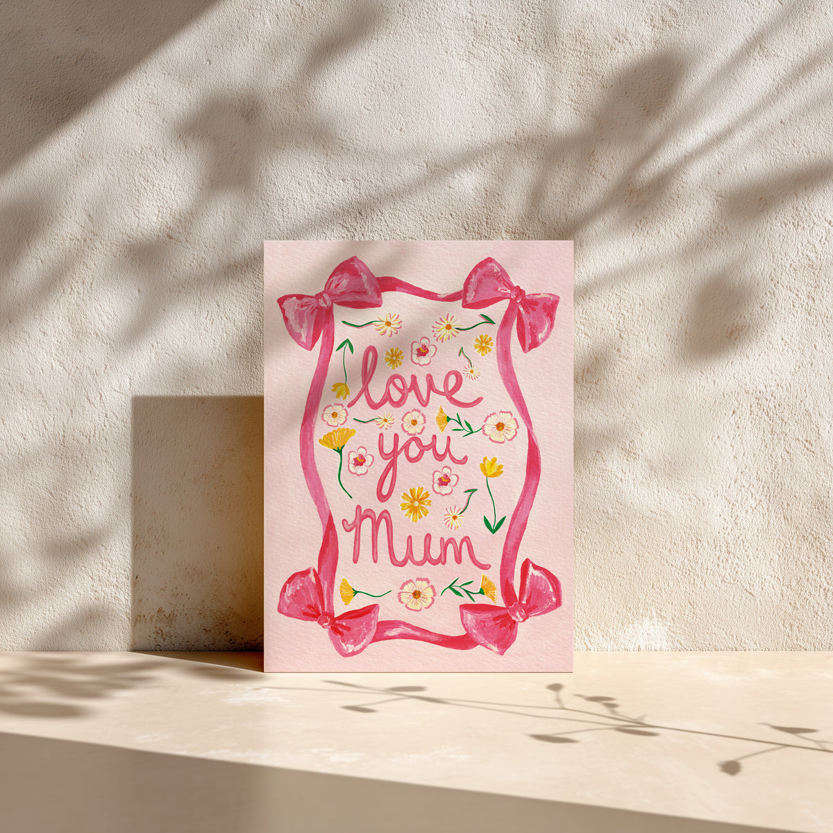 A pink greeting card with a watercolor-style pink border, bows, flowers, and the words 'love you Mum' in script, standing on a light surface against a textured wall with dappled shadows.