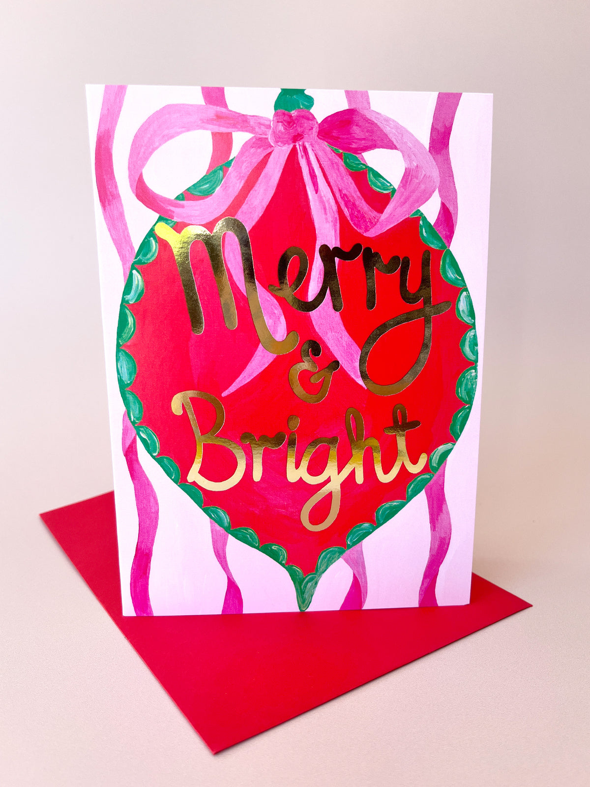 A festive Christmas greeting card with a red heart-shaped ornament design, pink ribbon bow, green border, and gold foil text reading 'Merry & Bright,' shown standing on a red envelope against a light beige background.
