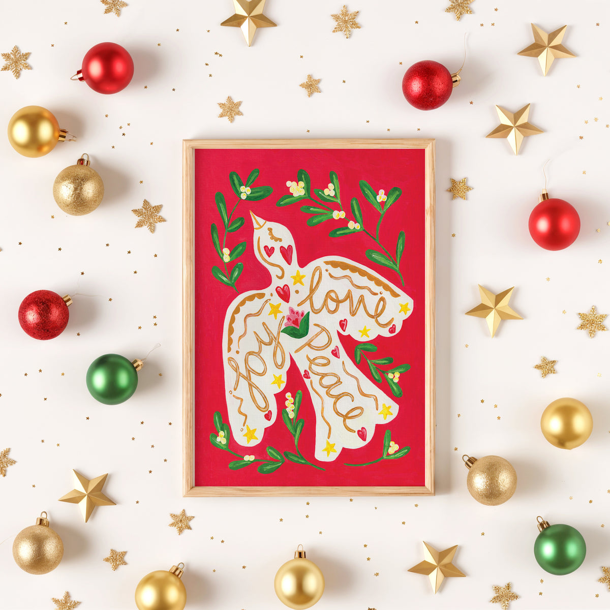 A framed Christmas art print featuring a dove with 'Joy, Love, Peace' written on its wings, surrounded by green holly leaves and small yellow flowers on a red background. The frame is placed on a white surface decorated with gold and red ornaments and gold star cutouts.