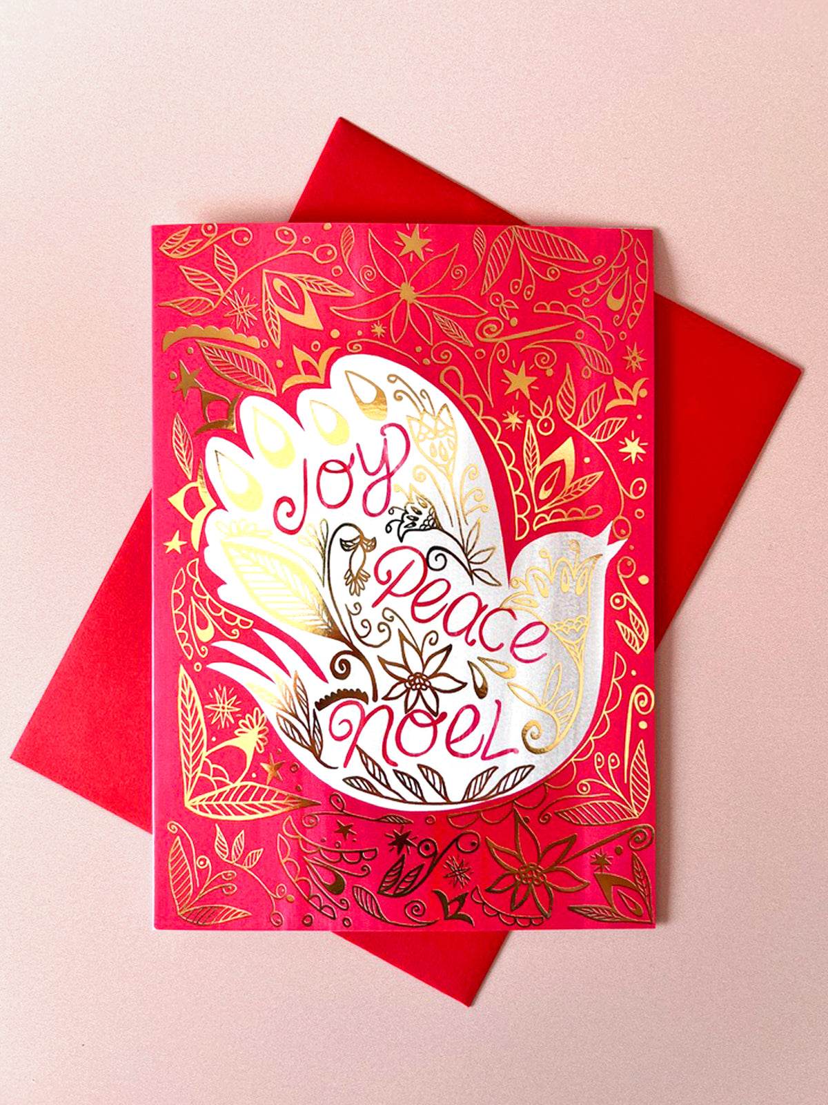 A festive red greeting card with gold floral patterns and a white dove illustration, featuring the words 'Joy Peace Noel' in stylized lettering, displayed on top of a red envelope against a light pink background.