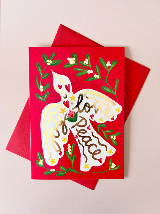A red greeting card featuring a white dove with the words 'love and peace' written inside, surrounded by green leaves and small red hearts, shown with a matching red envelope on a light beige background.