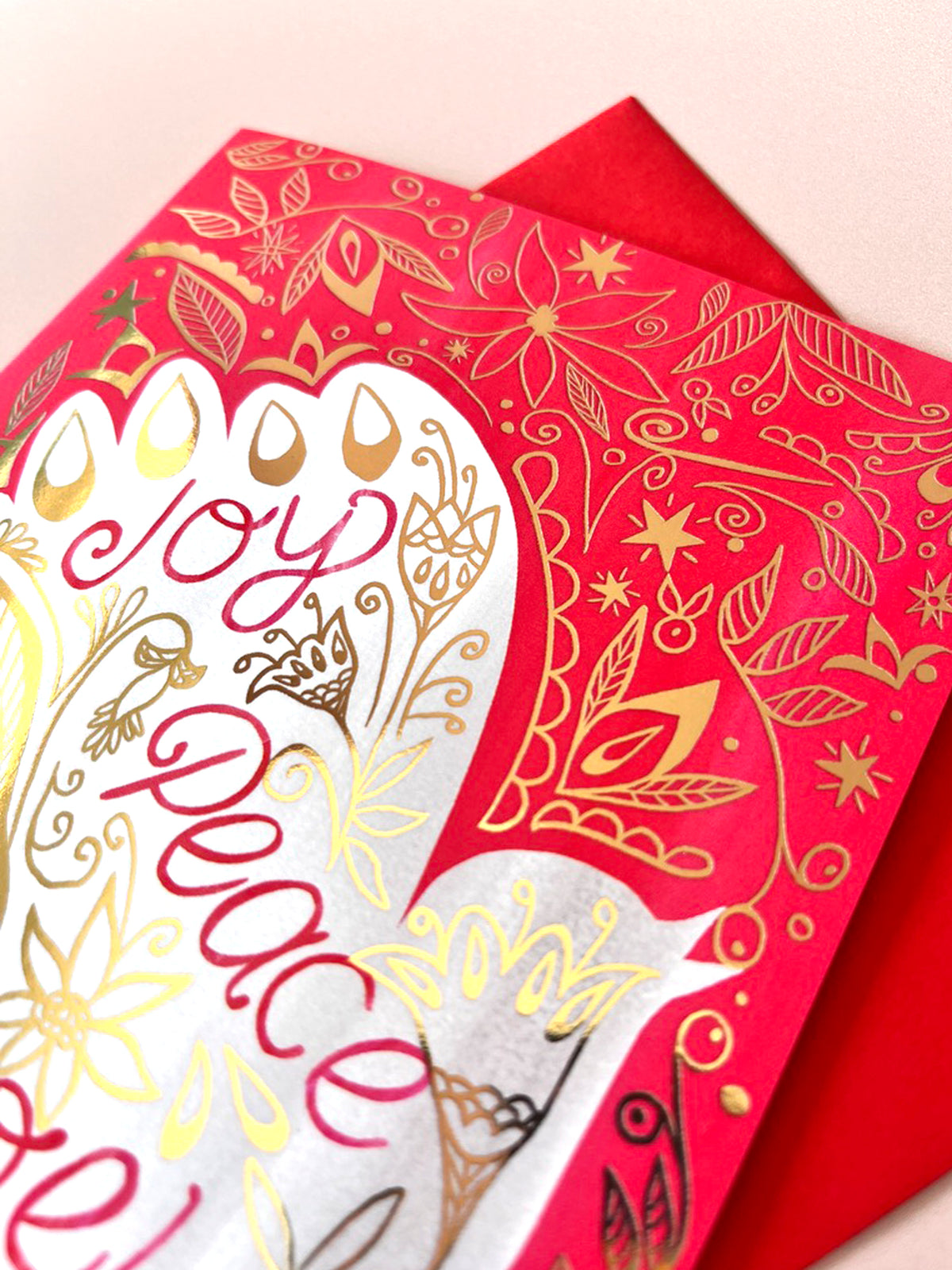 A close-up of a red Christmas greeting card with gold foil floral patterns and a white heart-shaped center featuring the words 'Joy Peace Love' in red and gold script, with a red envelope behind it.