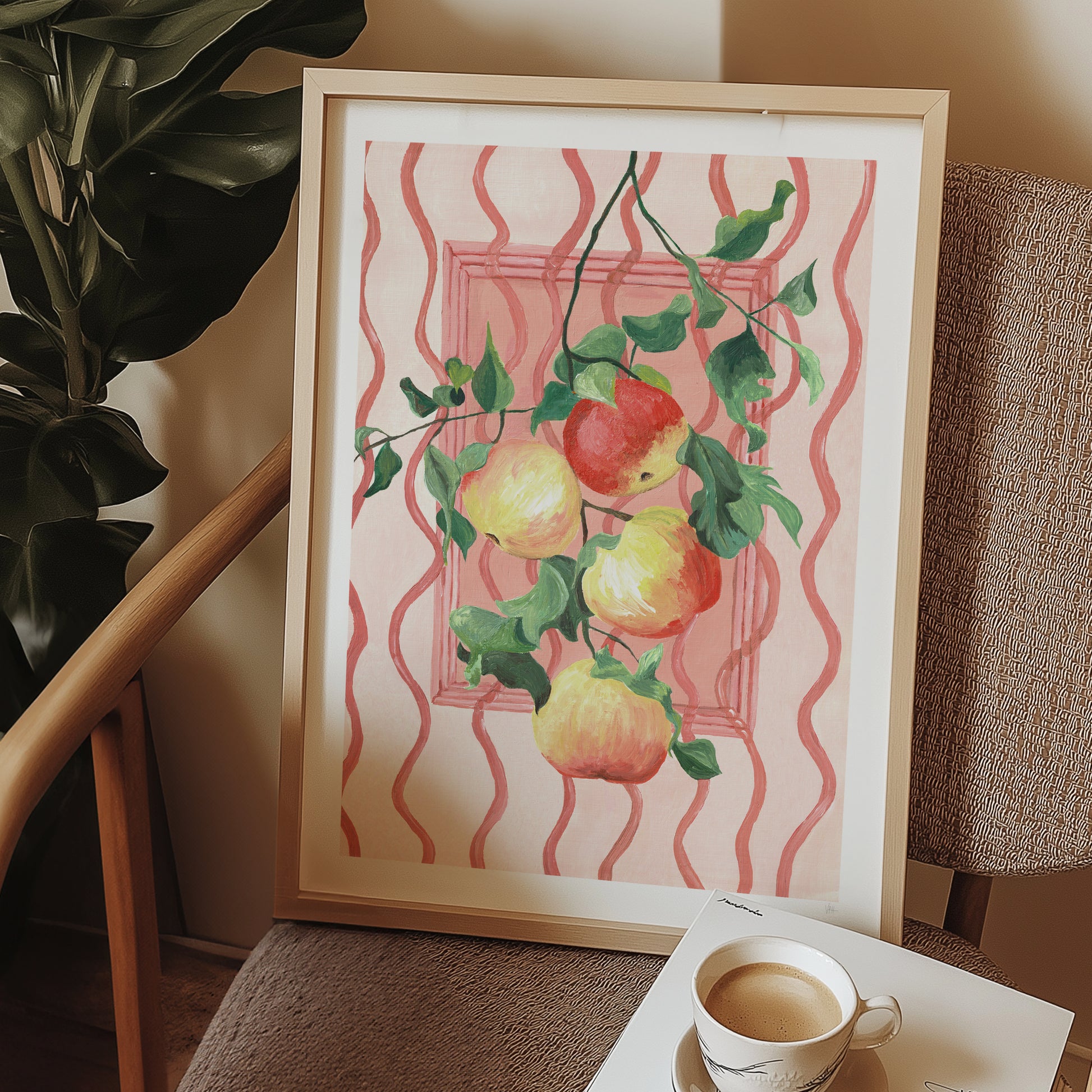Framed artwork of apples on a pink background next to a plant and a cup of coffee.