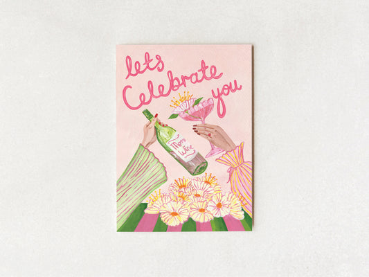 A square greeting card with a pink background and an illustration of hands clinking a wine bottle and a glass, surrounded by flowers, with the text 'let's Celebrate you' in pink script. The card is photographed flat against a light gray textured background.