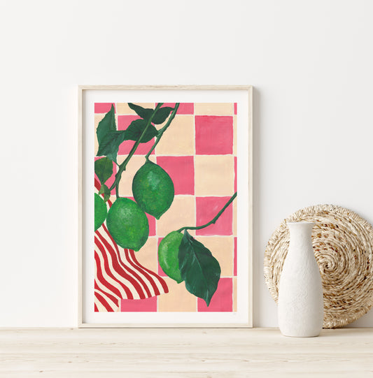 Framed artwork of limes on a pink and cream checkerboard pattern, placed on a wooden surface with a white wall background.