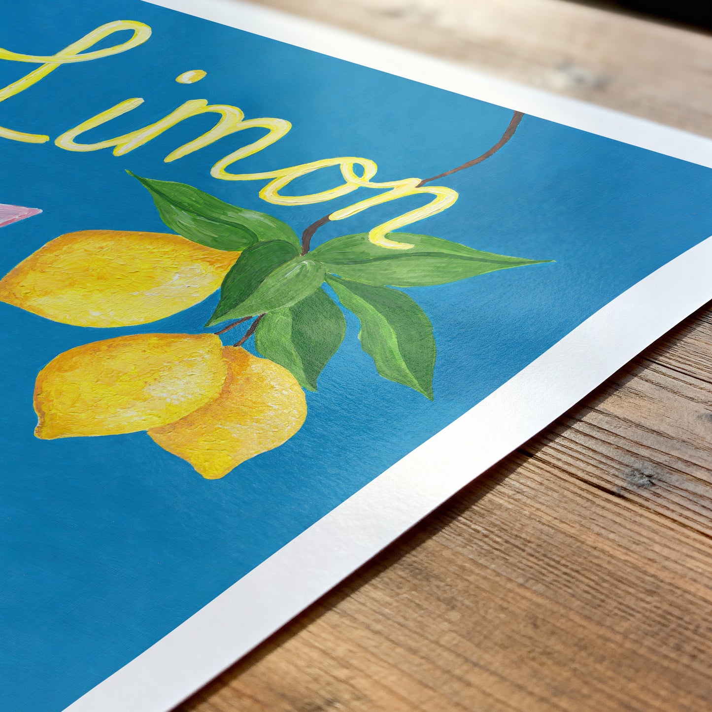 Art print with lemons and the word 'Limon' on a blue background, placed on a wooden surface.