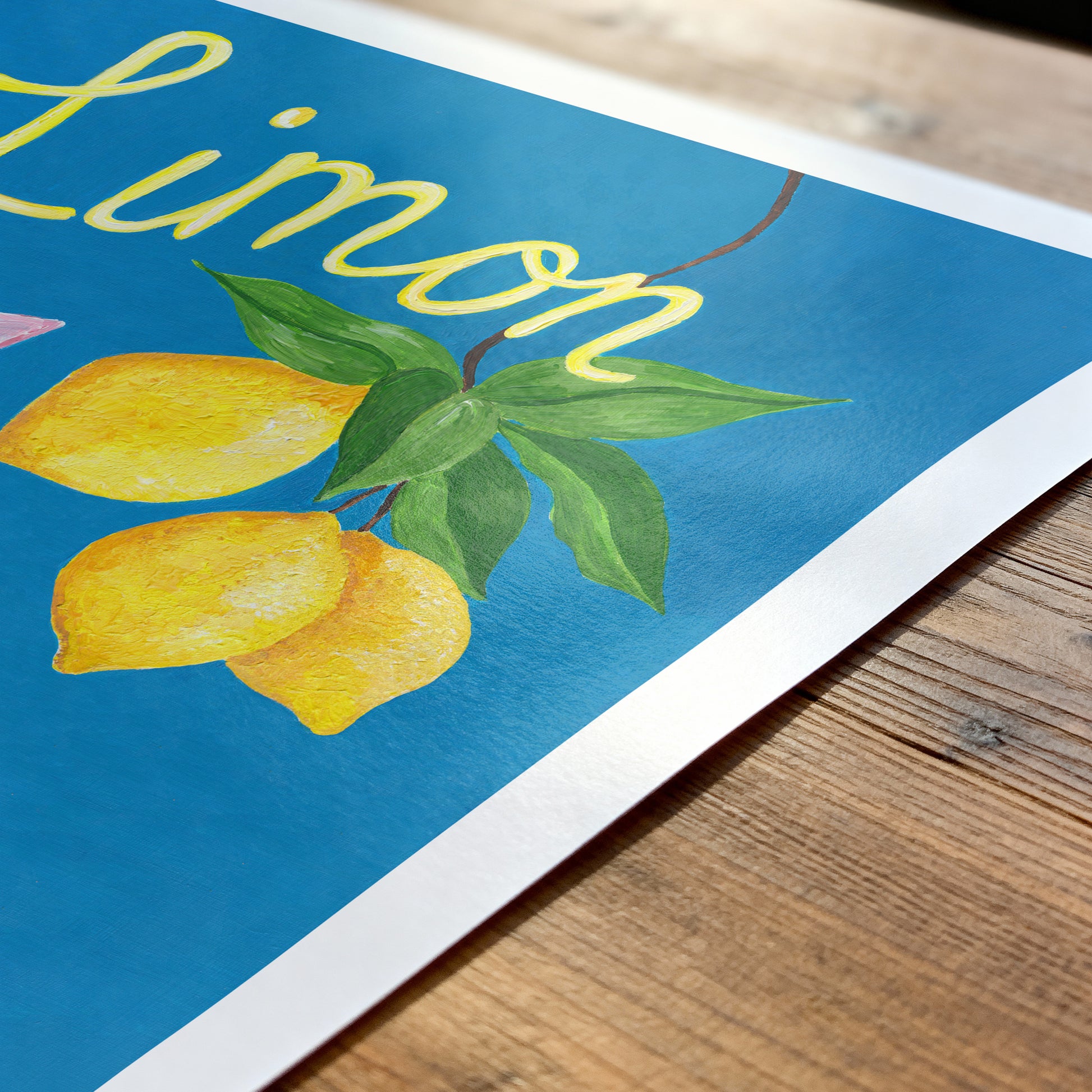 Art print with lemons and the word 'Limon' on a blue background, placed on a wooden surface.