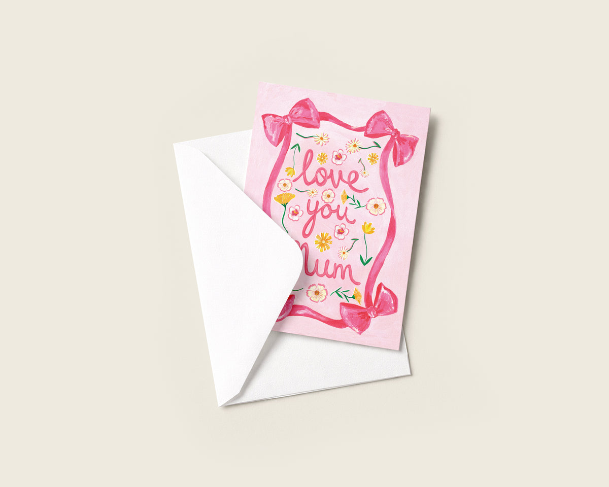 A pink greeting card with the words 'love you mum' in a floral border design, shown with a white envelope on a light beige background.