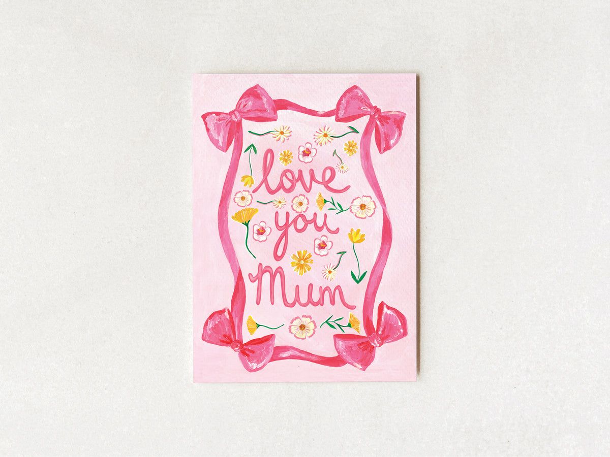 A pink greeting card with the words 'love you Mum' in cursive, decorated with pink bows, yellow and white flowers, and green leaves, displayed on a light textured background.