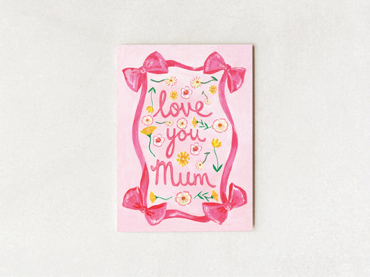 A pink greeting card with the words 'love you Mum' in cursive, decorated with pink bows, yellow and white flowers, and green leaves, displayed on a light textured background.