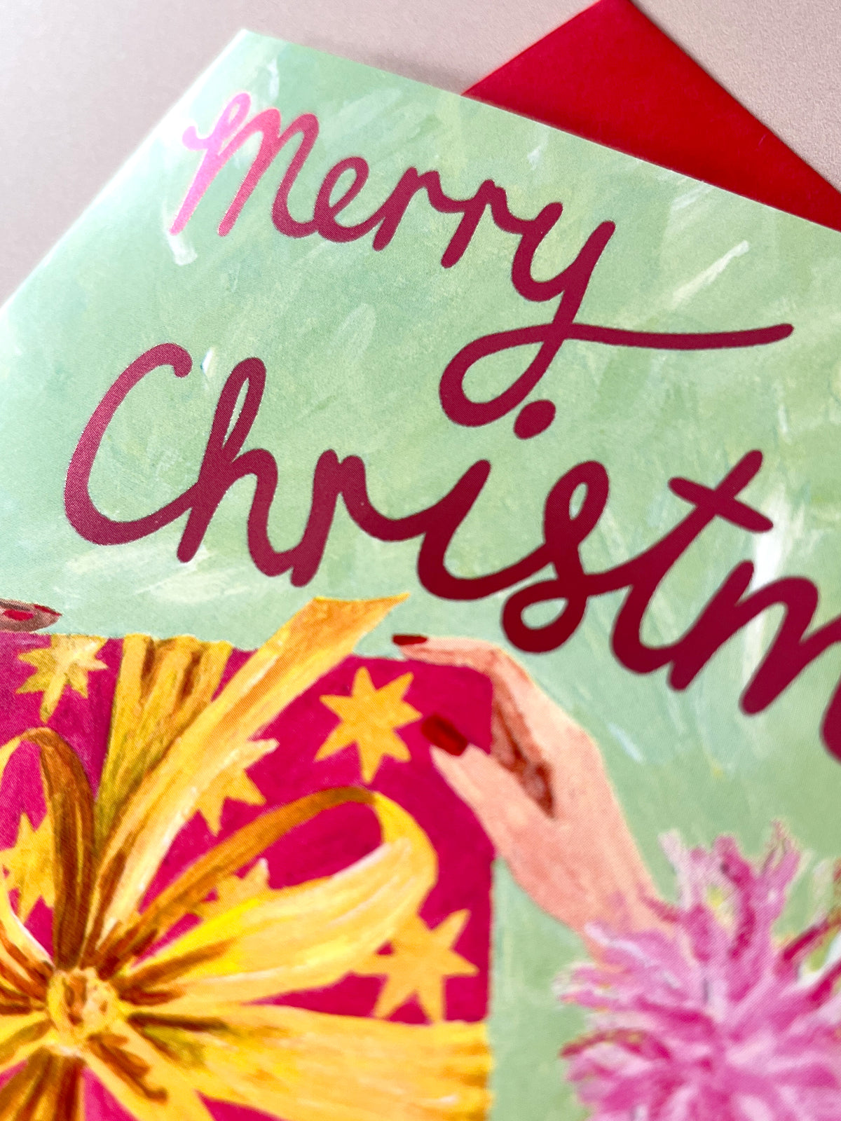 A close-up of a Christmas greeting card featuring the words 'Merry Christmas' in dark red script on a light green background, with painted illustrations of a gift box, stars, and a hand. A red envelope is partially visible behind the card.