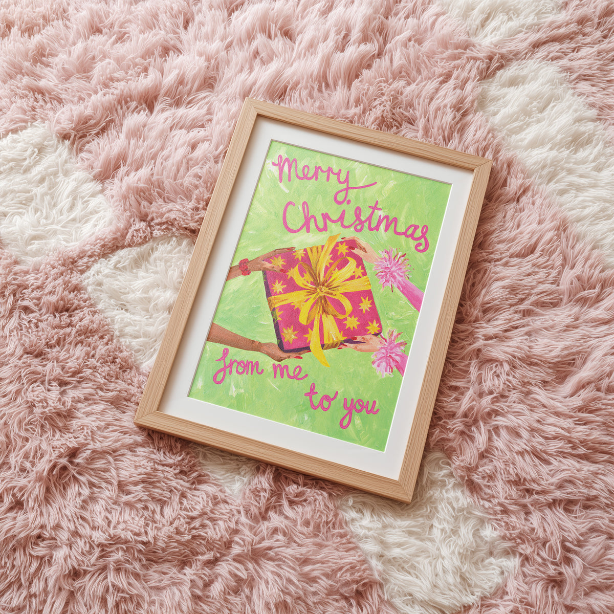 A framed Christmas greeting card art print featuring a pink gift box with a yellow ribbon on a light green background, with the text 'Merry Christmas from me to you' in pink script. The print is housed in a light natural wood frame and is placed on a fluffy pink and white rug.