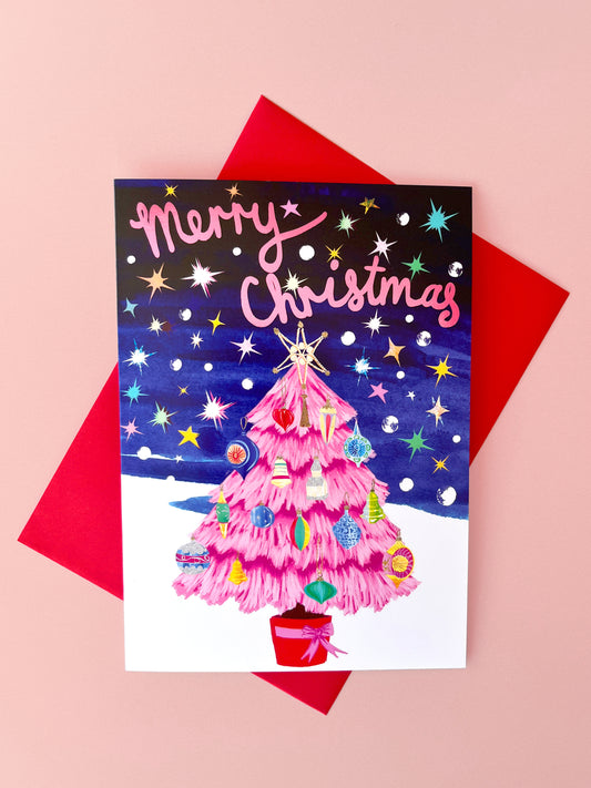 A Christmas greeting card featuring a pink tree with colorful ornaments under a starry night sky, placed on top of a red envelope against a pink background.