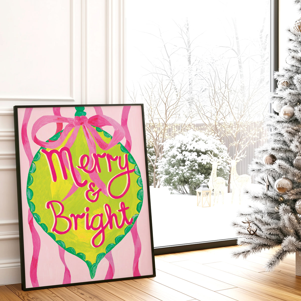 A framed Christmas art print leaning against a white wall on a wooden floor, showing a bright pink and lime green ornament design with the words 'Merry & Bright' in pink script. A snowy outdoor scene is visible through the window behind it, and part of a decorated Christmas tree stands on the right.