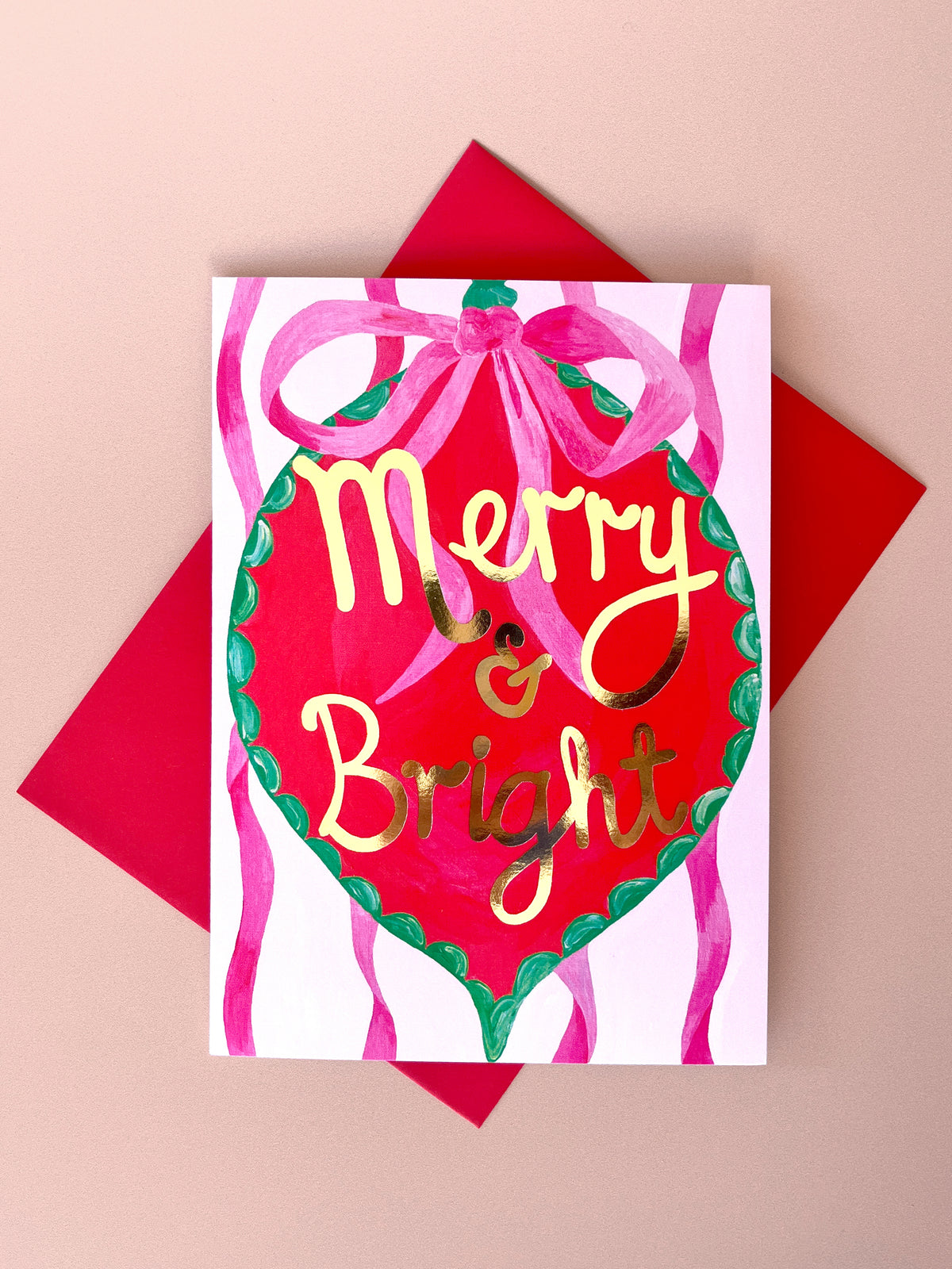 A festive greeting card featuring a red ornament illustration with pink ribbons and gold 'Merry & Bright' lettering, shown with a red envelope on a beige background.