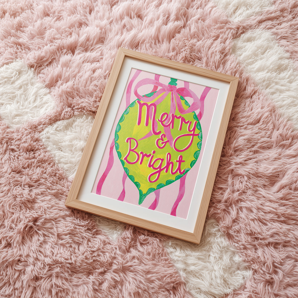 A framed art print featuring a colorful Christmas ornament illustration with the words 'Merry & Bright' in pink script, placed on a fluffy pink and white rug.