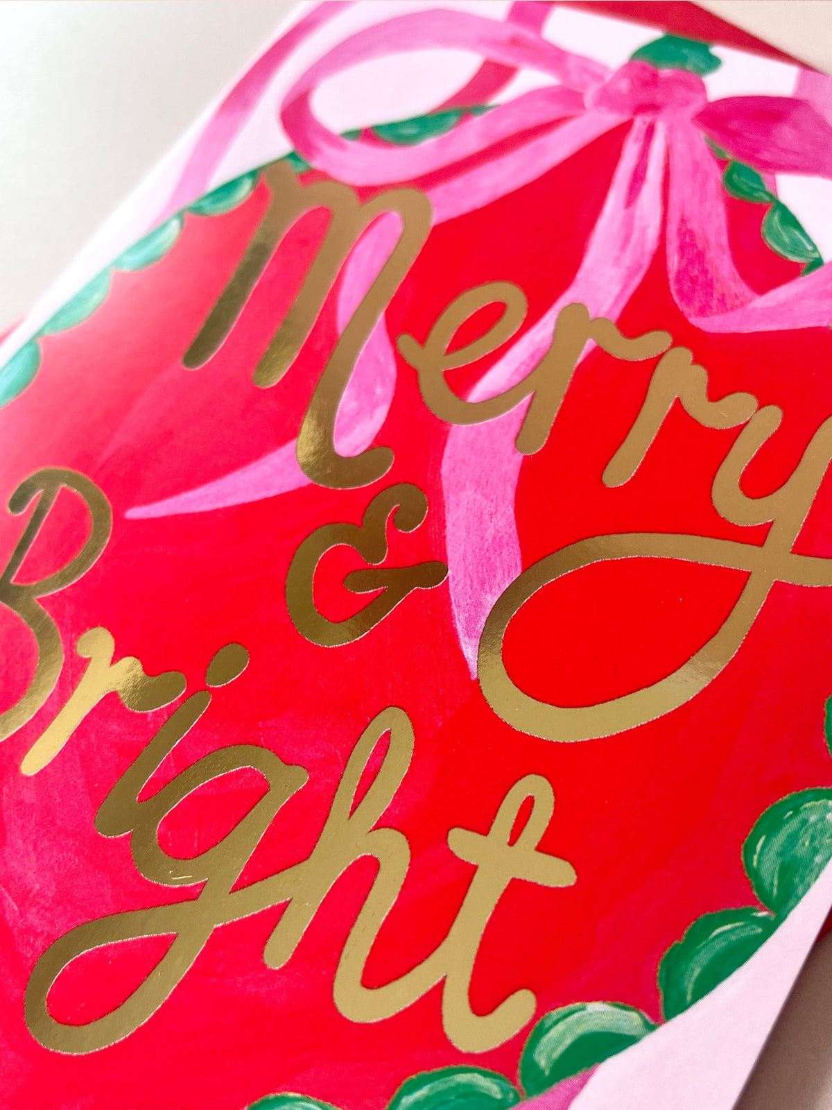 A close-up of a red Christmas greeting card featuring metallic gold foil script that reads 'Merry & Bright' beneath a pink ribbon bow, with green holly leaves around the border.