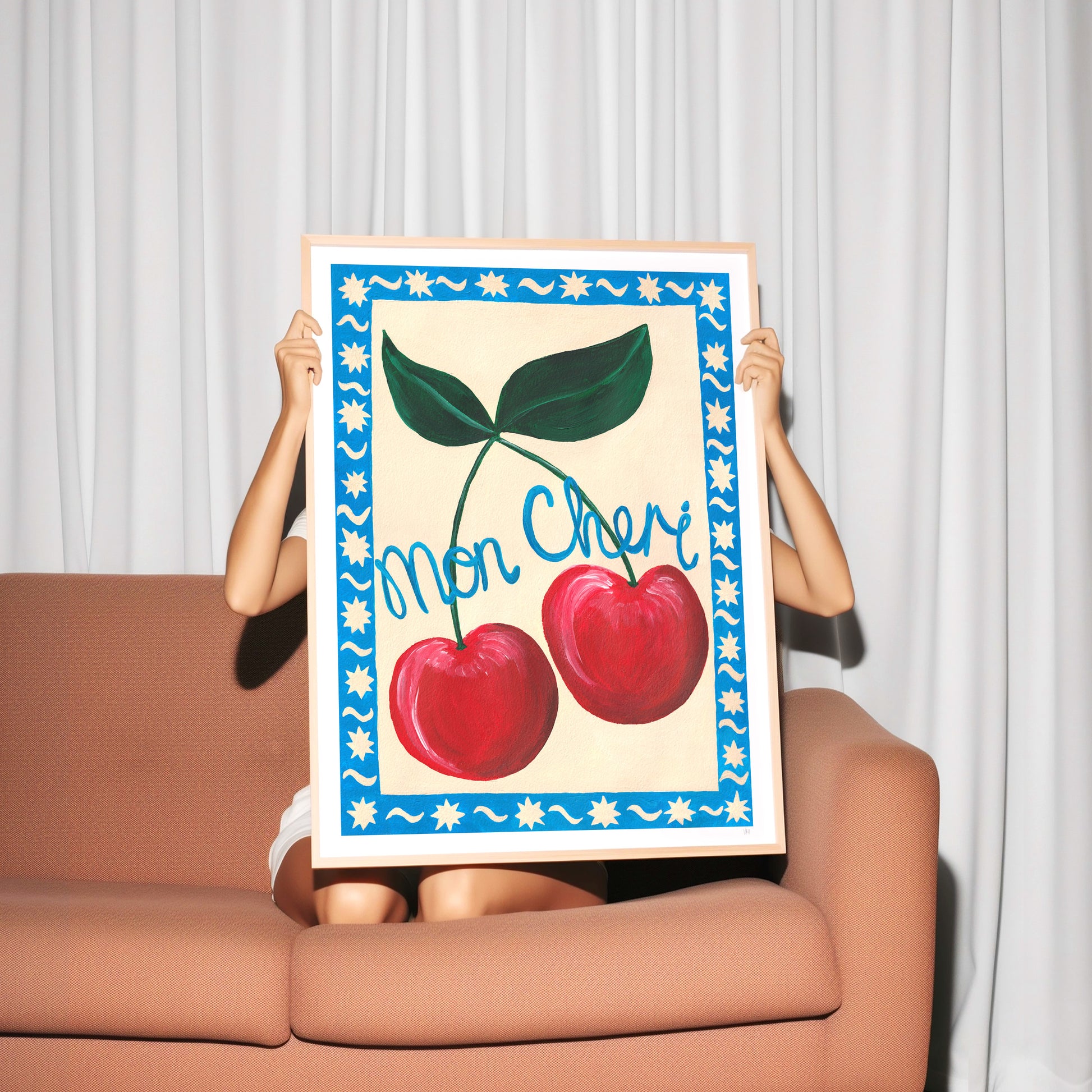 Person holding a framed artwork with cherries and 'Mon Cheri' text against a white curtain background.