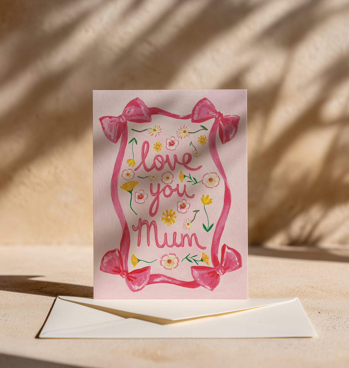 A pink greeting card with a watercolor-style floral border and the words 'love you Mum' in pink script, displayed upright on a beige surface with an envelope in front and warm shadow patterns in the background.