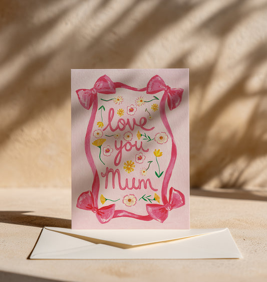 A pink greeting card with a watercolor-style floral border and the words 'love you Mum' in pink script, displayed upright on a beige surface with an envelope in front and warm shadow patterns in the background.