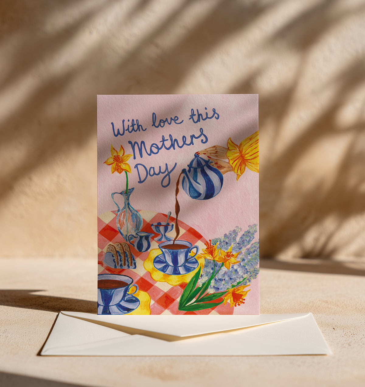 A square Mother's Day greeting card with a pastel illustration of a tea-time scene and the text 'With love this Mothers Day,' displayed upright on a cream envelope against a warm beige background with soft leaf shadows.