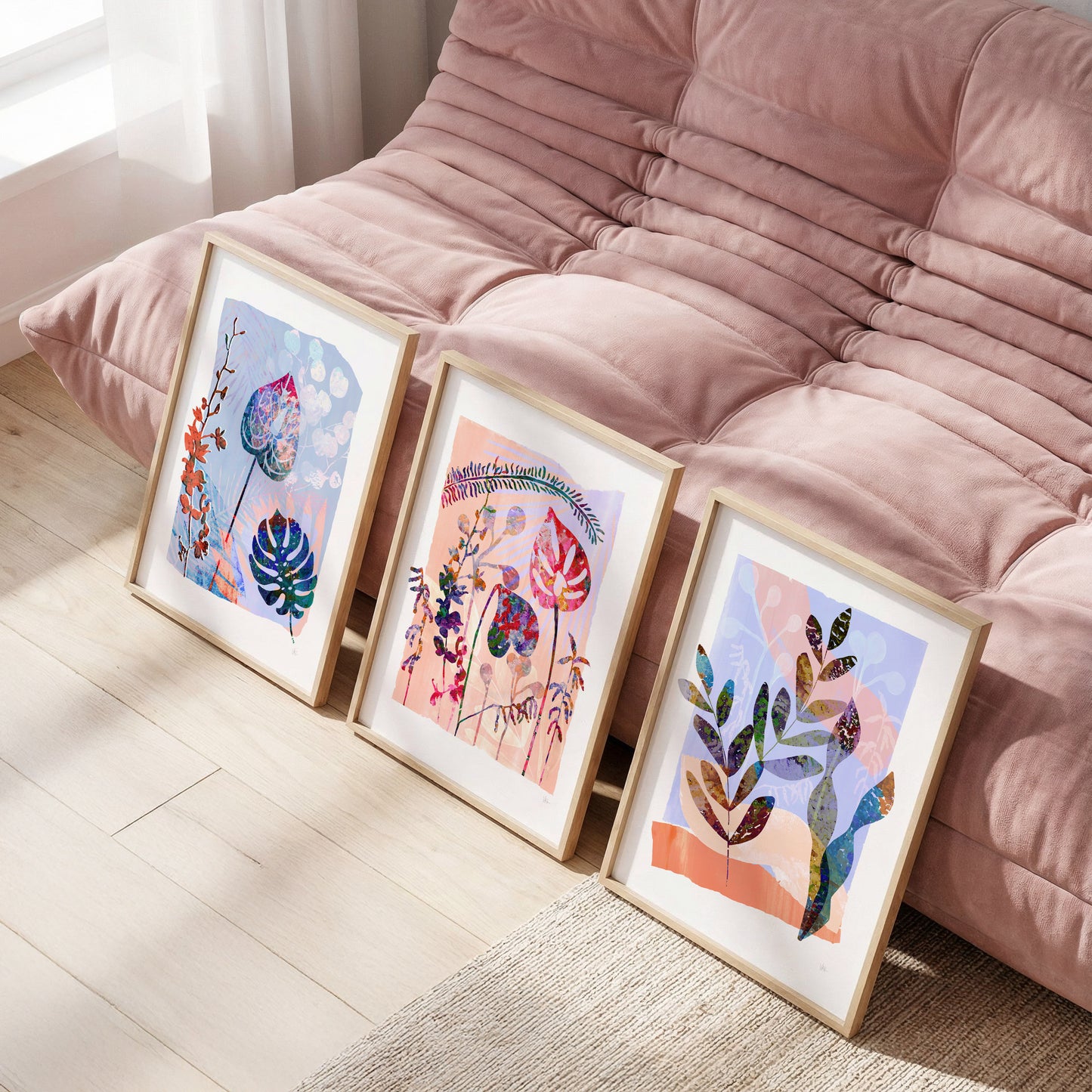 Three framed artworks on a wooden floor in front of a pink sofa.
