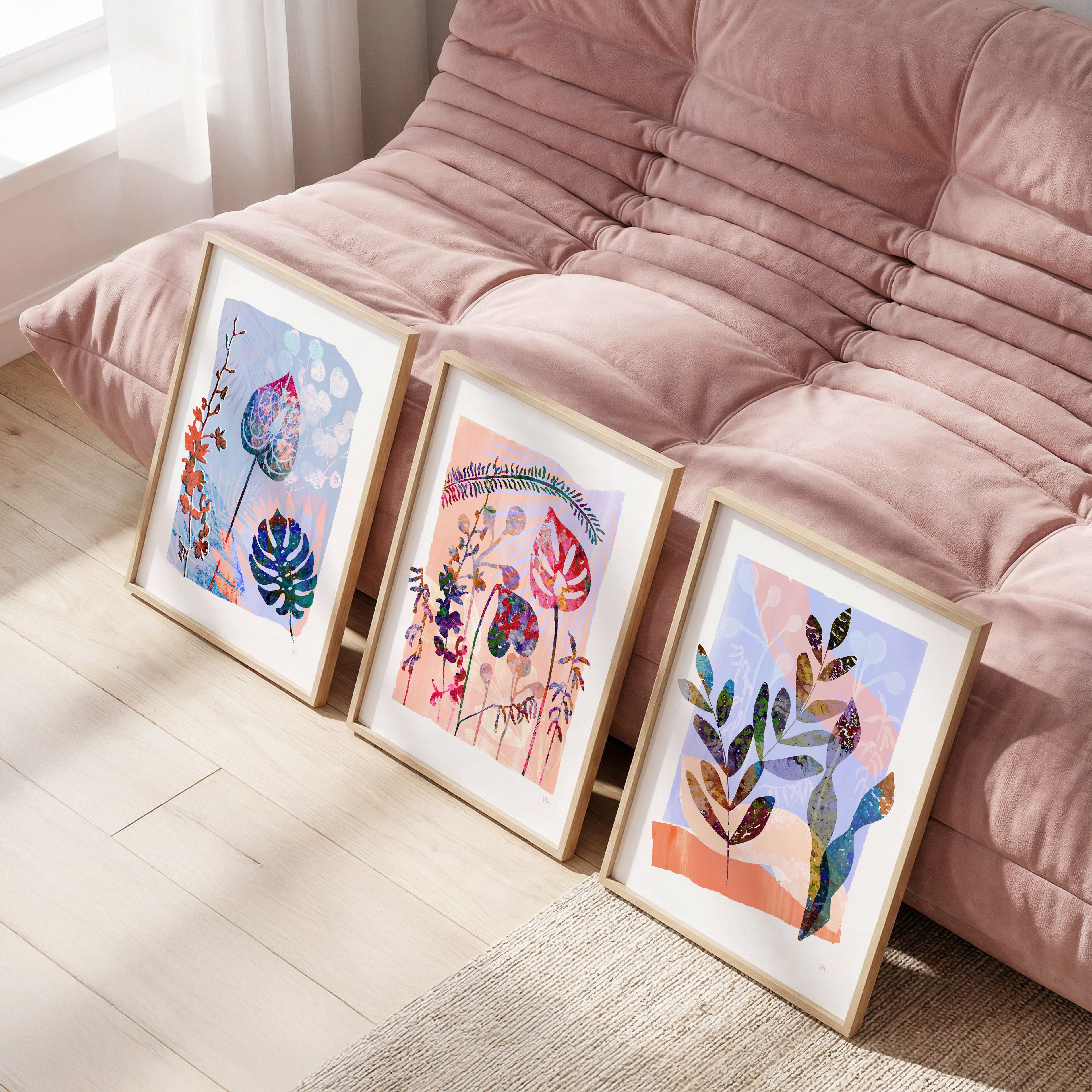 Three framed artworks on a wooden floor in front of a pink sofa.