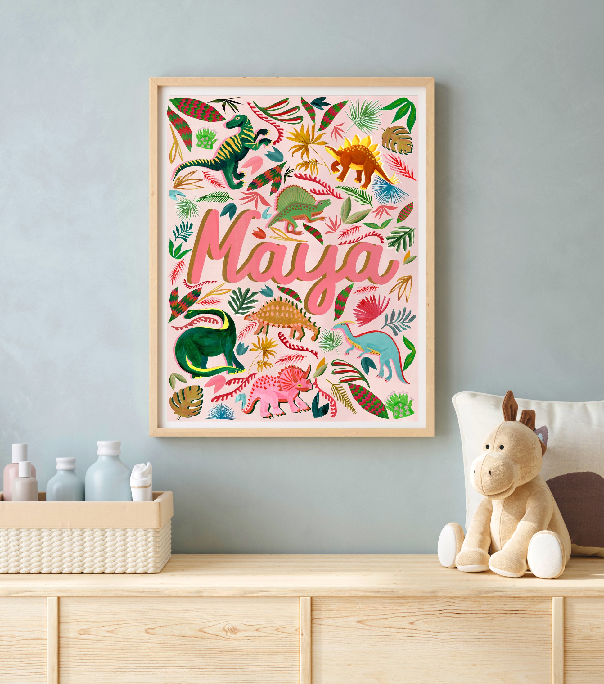 Framed children's art print with colorful dinosaurs and the name 'Maya' on a wall above a wooden shelf.