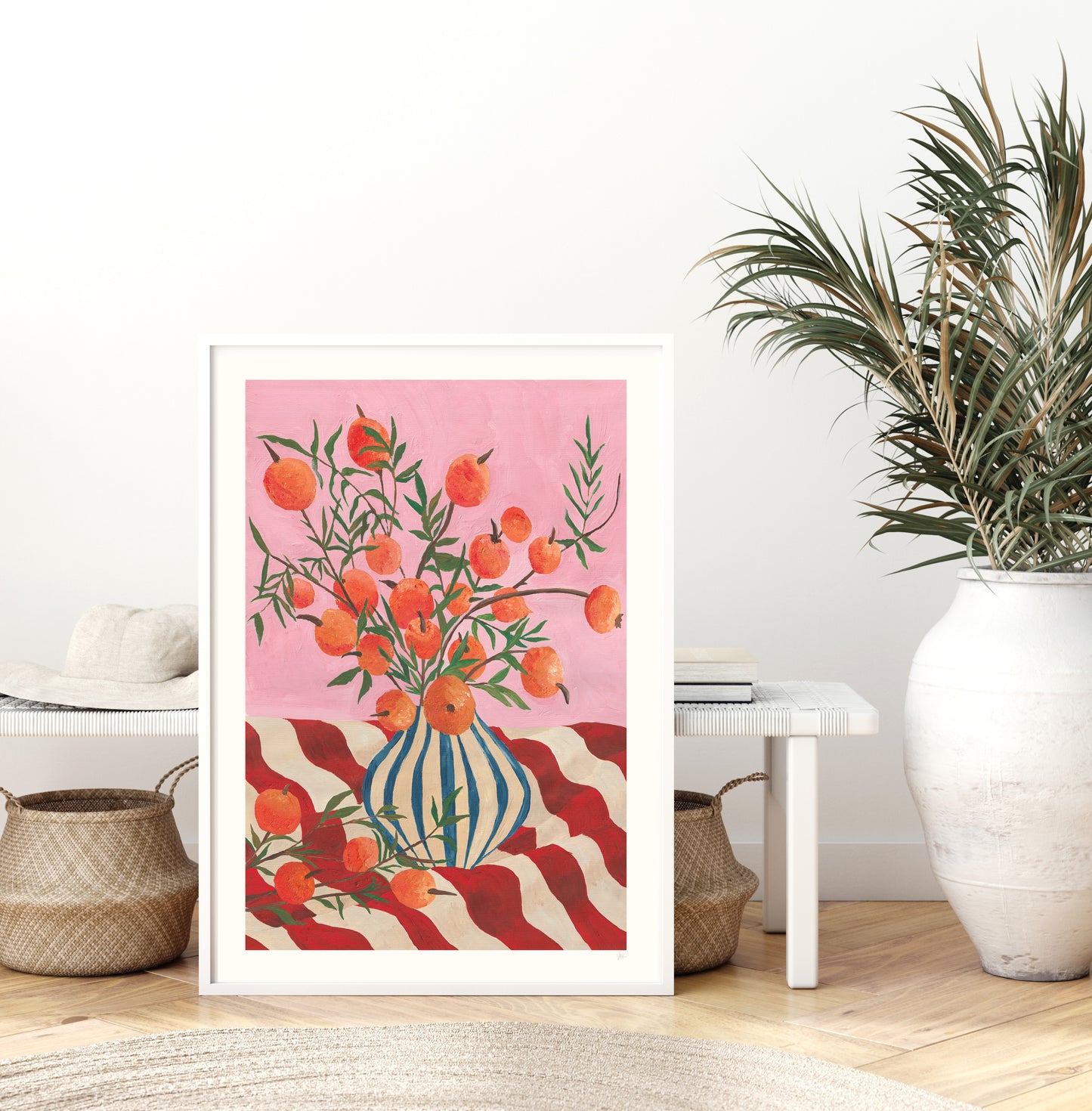 Framed artwork with a fruit in a vase design on a pink background in a room with a plant and furniture.