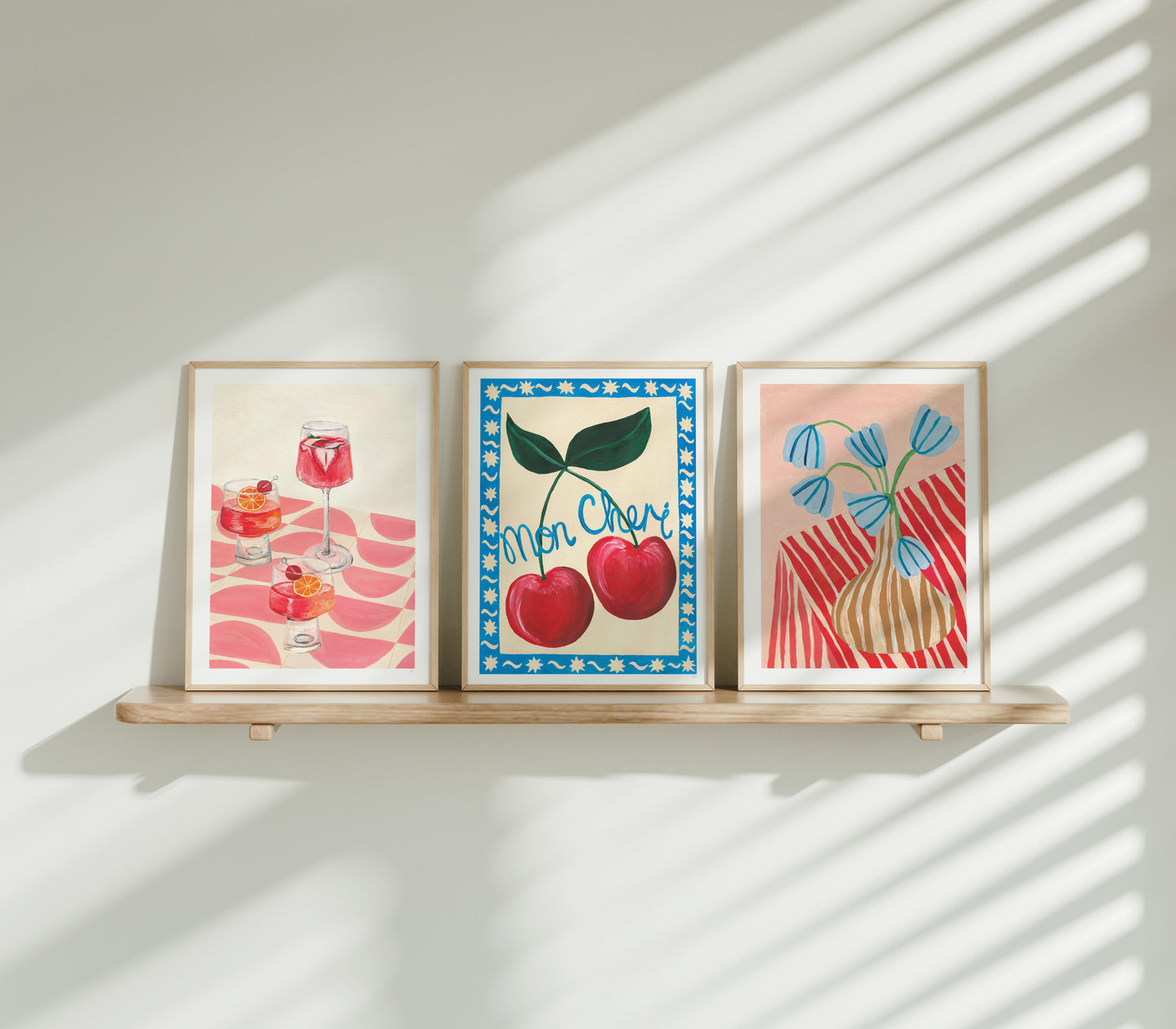 Three framed artworks on a wooden shelf with sunlight casting shadows on a white wall.