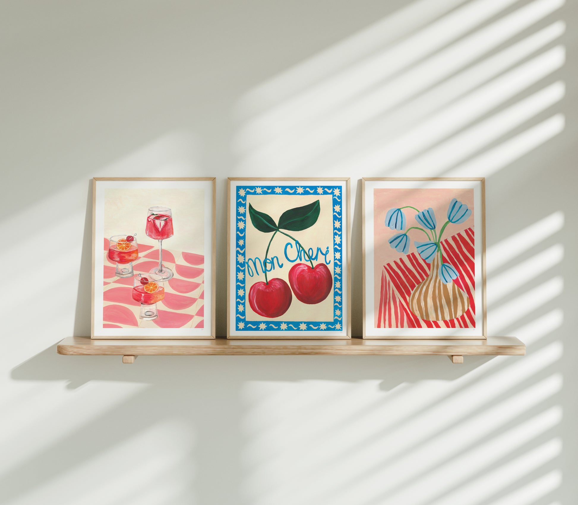 Three framed artworks on a wooden shelf with sunlight casting shadows on a white wall.