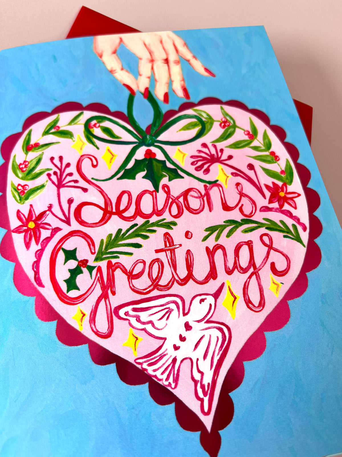 A square canvas painting featuring a large pink heart with scalloped magenta edges, decorated with red cursive text reading 'Seasons Greetings,' green holly leaves and vines, yellow stars, and a white dove near the bottom. A hand with red nail polish hangs from a green bow at the top.