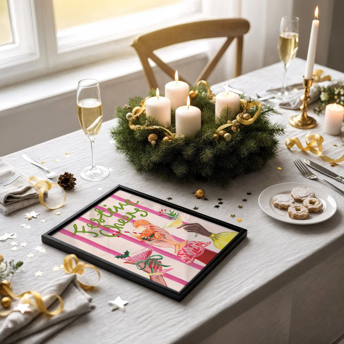 A framed art print with a colorful illustration and the text 'Letting Go' is displayed on a decorated dining table. The table setting includes a green pine wreath with white candles, gold ribbons, champagne flutes, and cookies on a plate.