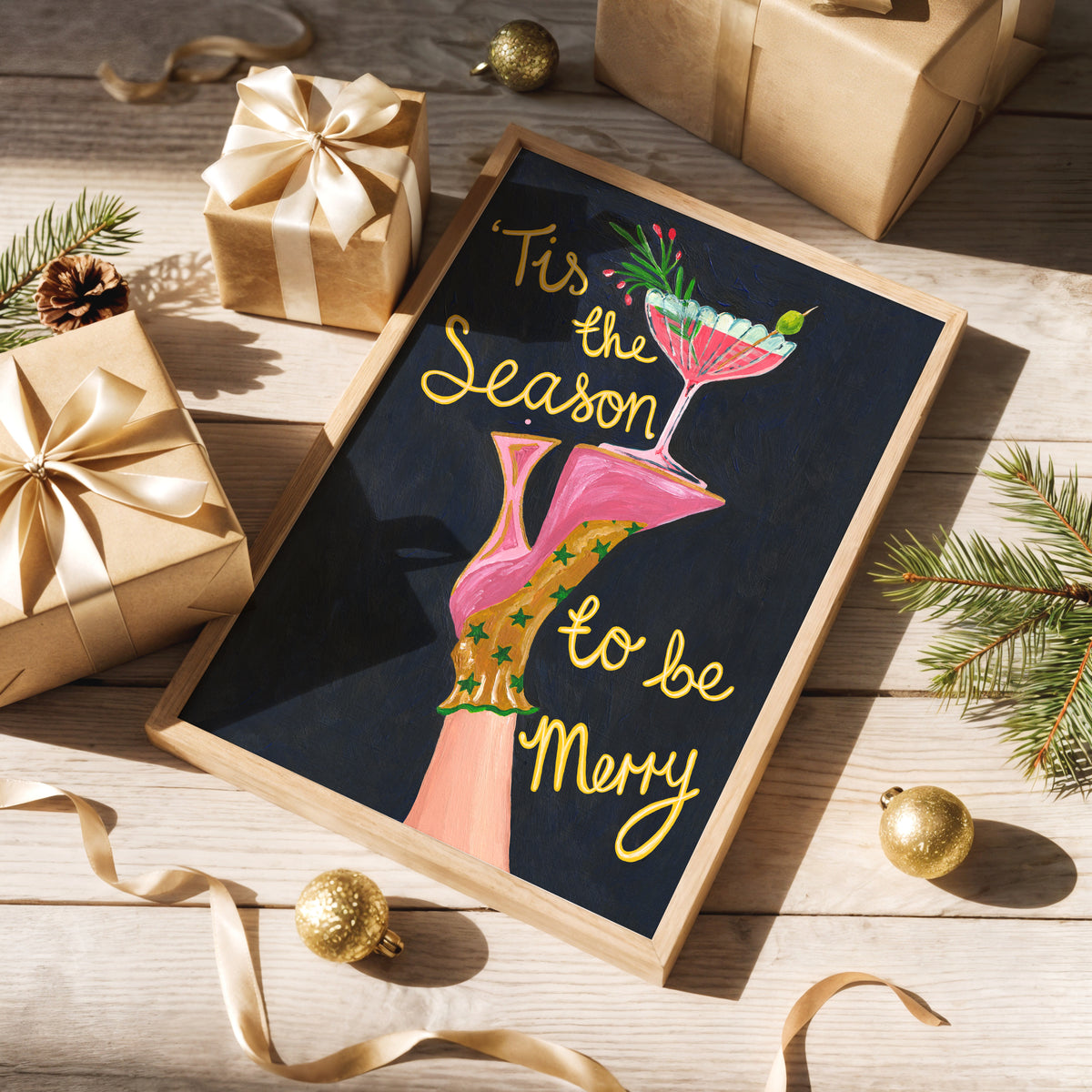 A framed holiday art print with the text "Tis the Season to be Merry" and an illustration of a pink high-heel boot holding a cocktail glass, styled on a wooden tabletop with wrapped gifts, pine branches, and gold ornaments.