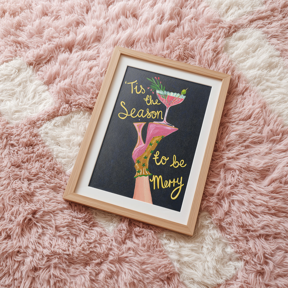 A framed art print featuring a black background with the phrase 'Tis the Season to be Merry' in gold script, alongside an illustration of pink high heels and a cocktail glass, displayed on a pink and white shag rug.