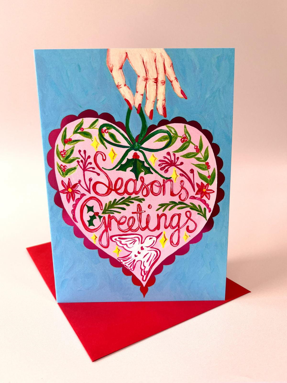 A square greeting card with a hand holding it from the top, featuring a large pink heart illustration with the words 'Season's Greetings' and holiday floral details, set against a light blue background with a red envelope underneath on a pale pink surface.
