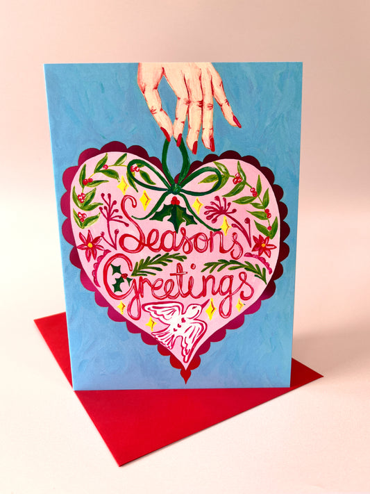 A square greeting card with a hand holding it from the top, featuring a large pink heart illustration with the words 'Season's Greetings' and holiday floral details, set against a light blue background with a red envelope underneath on a pale pink surface.